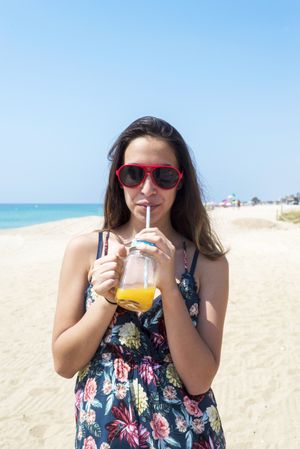 A dark haired woman in dress and sunglasses, holding out a delicious juice drink in her hand