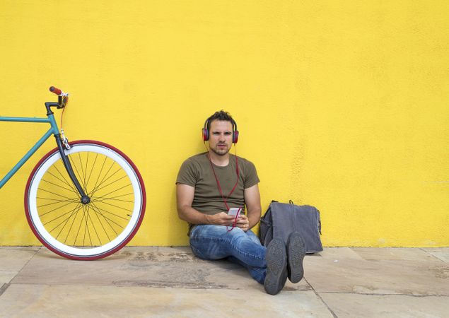 Male relaxing in front of yellow wall next to bike listening to music on smartphone