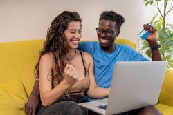 An excited diverse couple celebrating a successful online purchase with a credit card and laptop at home.