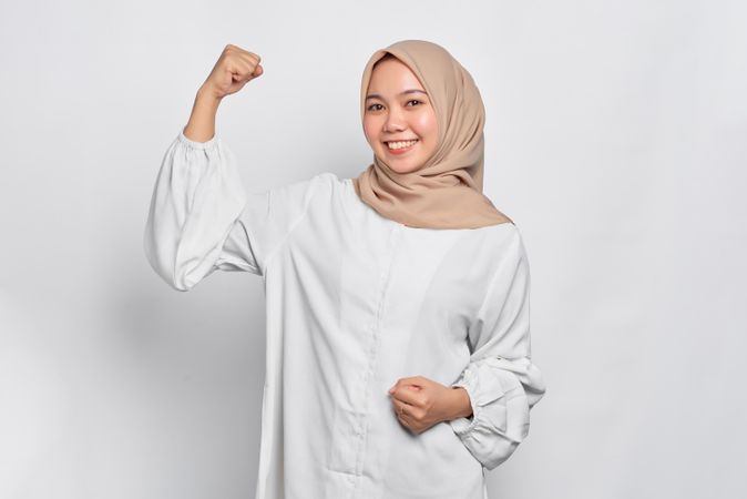 Muslim woman in headscarf and light blouse and smiling and flexing her biceps