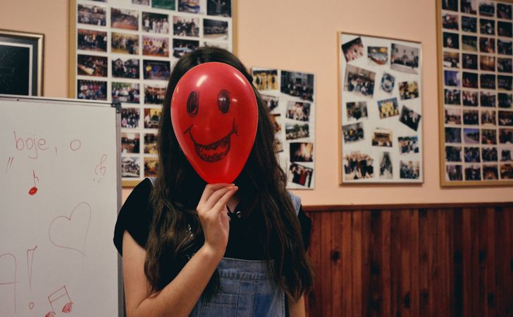 Teenage girl in dungaree hiding her face with red balloon