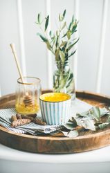 Wood tray with latte coffee, honey dipper, and vase with olive leaves