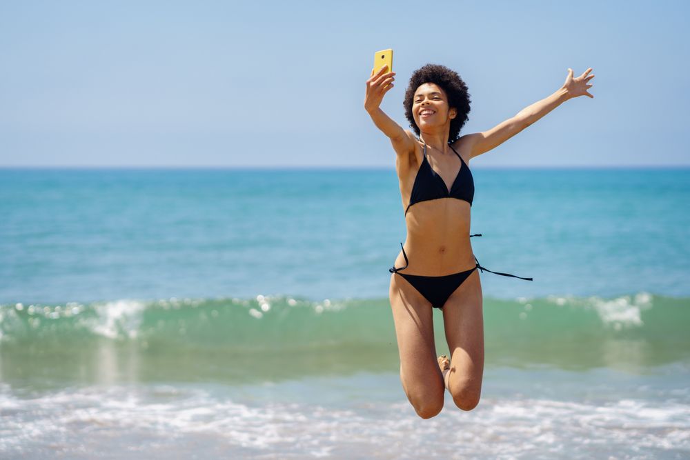 Woman in bikini happily jumping in front of the ocean and taking a selfie