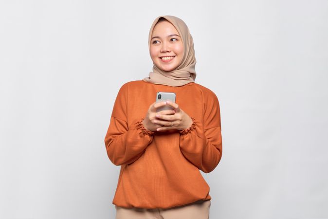 Smiling Muslim woman in headscarf and orange shirt with phone and looking away