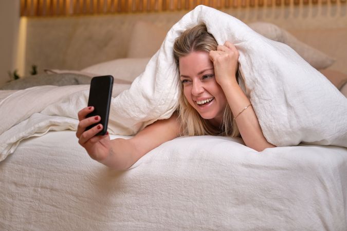 Young female taking selfie on smartphone while lying on bed