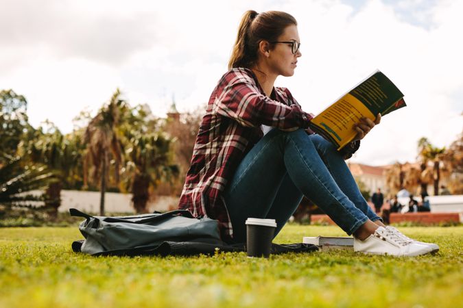 Side view of young woman sitting outdoors at college campus reading a book
