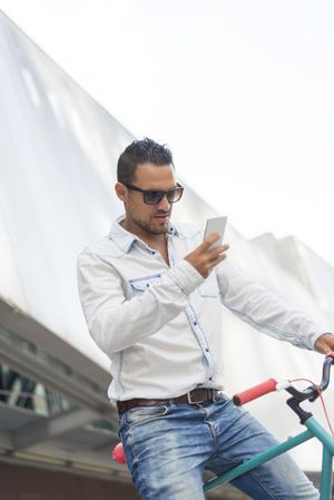 Man in denim sitting on bike looking at smartphone in front of building