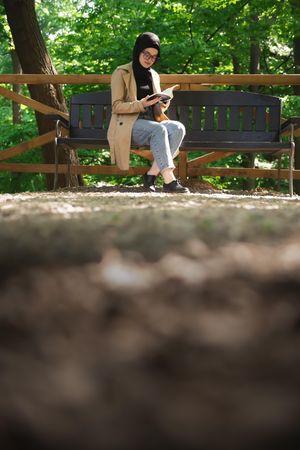 Woman in headscarf and trench coat reading on park bench