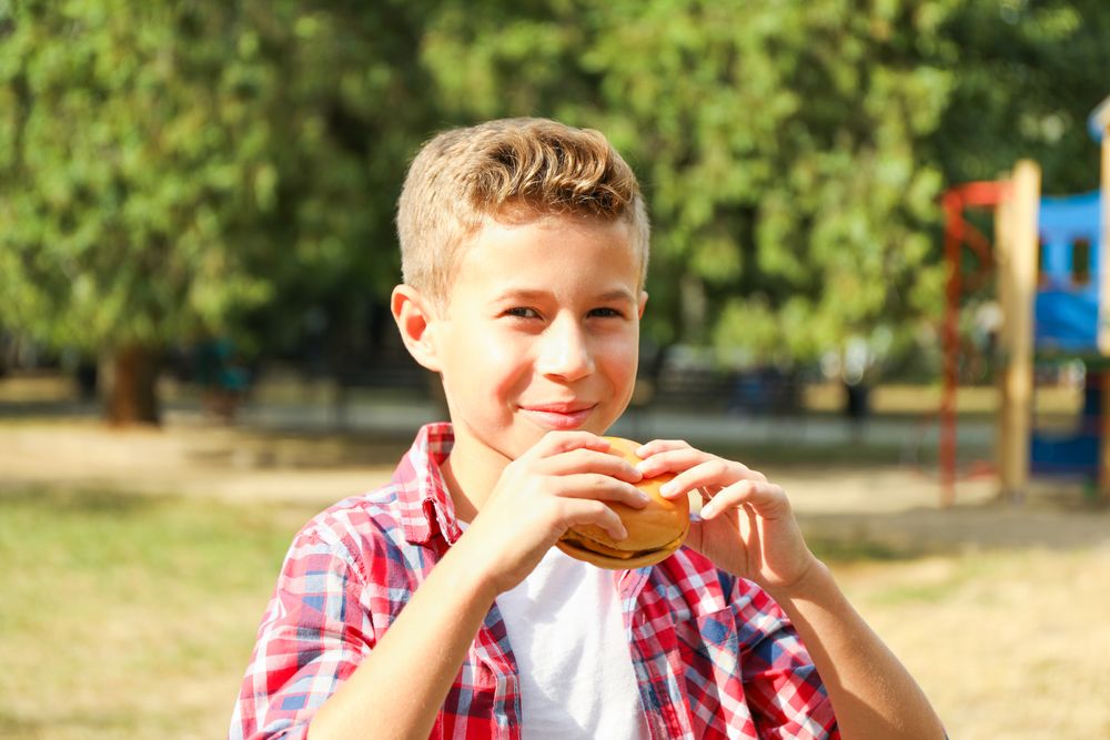 Boy with burger against blurred playground background, space for text