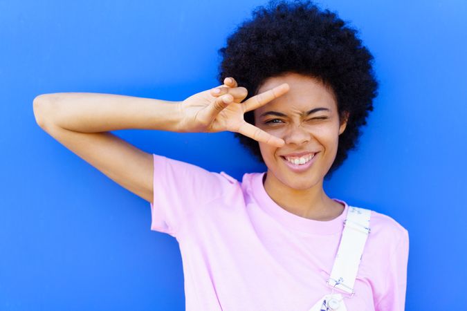 Woman in pink t-shirt making peace sign over eye against blue background