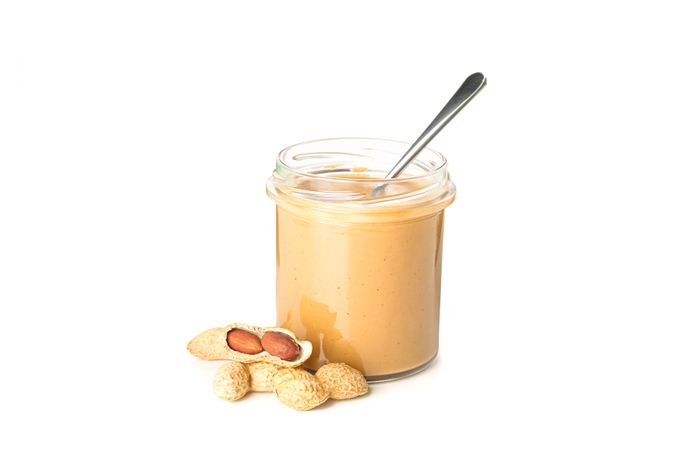 Creamy peanut butter in glass jar, peanut and spoon isolated on light background. A traditional product of American cuisine