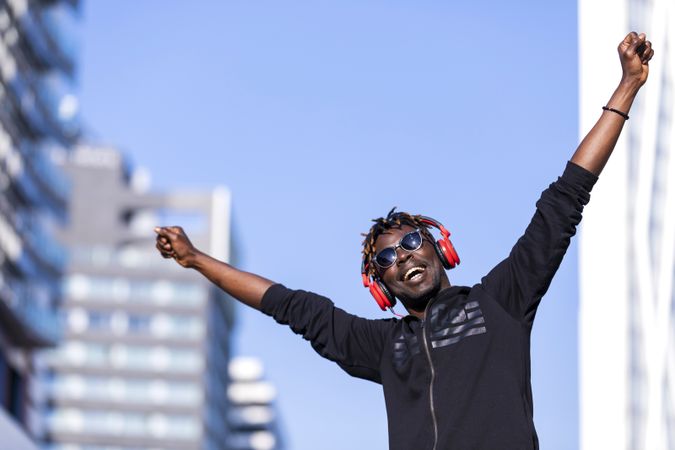 Joyful Black man wearing headphones & sunglasses standing on the street listening to music