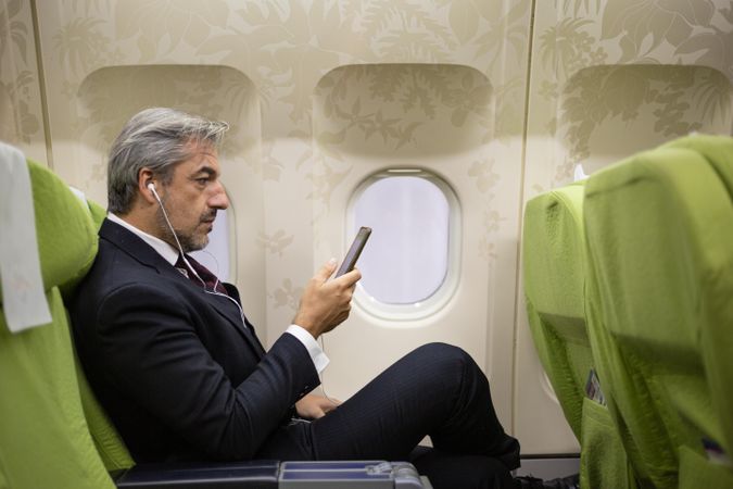 Man sitting in window seat watching phone screen with ear buds in flight