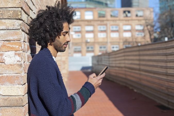 Side view of man looking down at his smartphone while leaning on a brick wall outdoors on sunny day