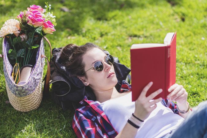 Side view of a hipster happy woman lying on grass in sunny day at park while reading a red book