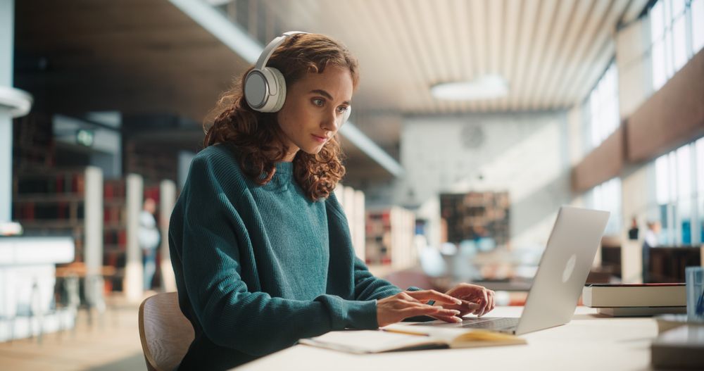 Concentrated Young Woman Studying in Modern Library, Wearing Headphones and Using Laptop for Research.