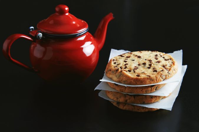 Shiny red tea pot on dark table with pile of cookies