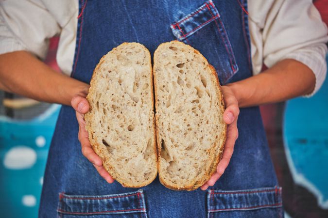 Photo detail of female hands holding an artisan bread broken in half with a blue kitchen apron.