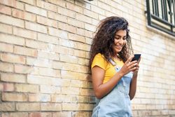 Smiling Arab woman texting with her smart phone leaning on brick wall, copy space