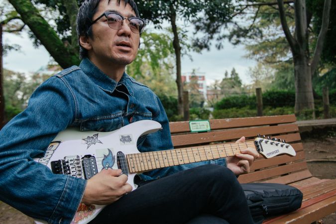 Man wearing denim jacket sitting on wooden bench and playing electric guitar in the park