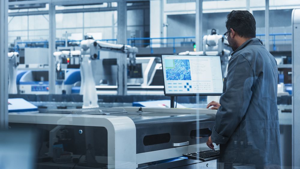 Indian Male Assembly Line Worker Using Soldering Jet Printer For Production Of Printed Circuit Boards At Autonomous Factory With Robotic Arms.