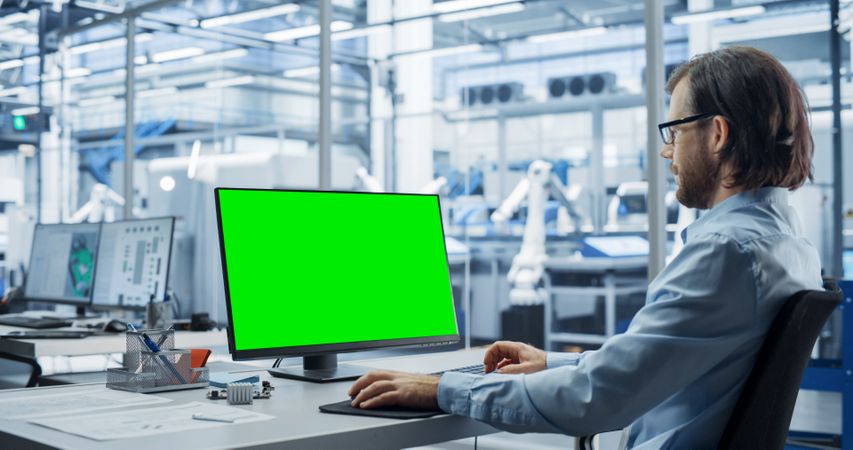 Robotics Engineer Working on a Desktop Computer with Green Screen Chromakey Mock Up Display in a Factory Office. Technological Research and Development Facility with Autonomous Assembly Conveyor