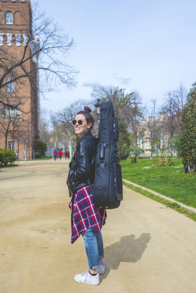 Side view of a young smiling hipster woman walking in a park in sunny day while carrying a guitar on back