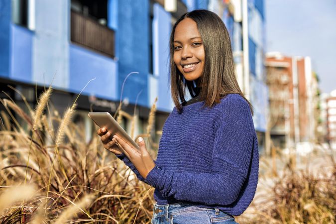 Happy female in sweater standing outside blue building in long grass with tablet