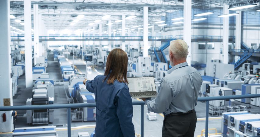 Experienced Male and Female Engineers Standing on a Platform, Using Laptop Computer and Discussing Production at a Modern Automated Electronics Manufacture with AI Support