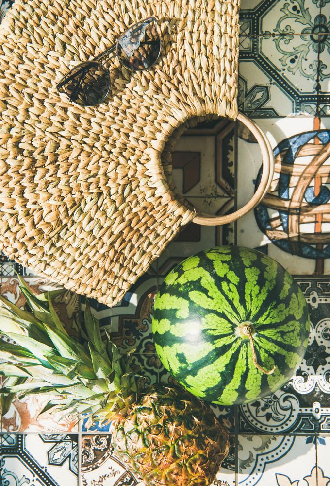 Thatched bag laying on pattered tiles with sunglasses, watermelon and pineapple