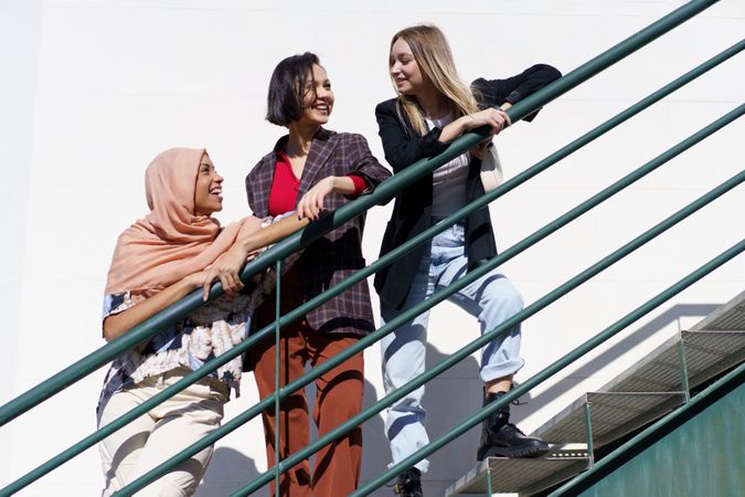 Low angle of trendy multi-ethnic women friends, in stylish outfit and hijab on outdoor stairs