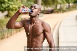 Fit male with shirt off drinking from a water bottle in an outdoor park 47Akz0