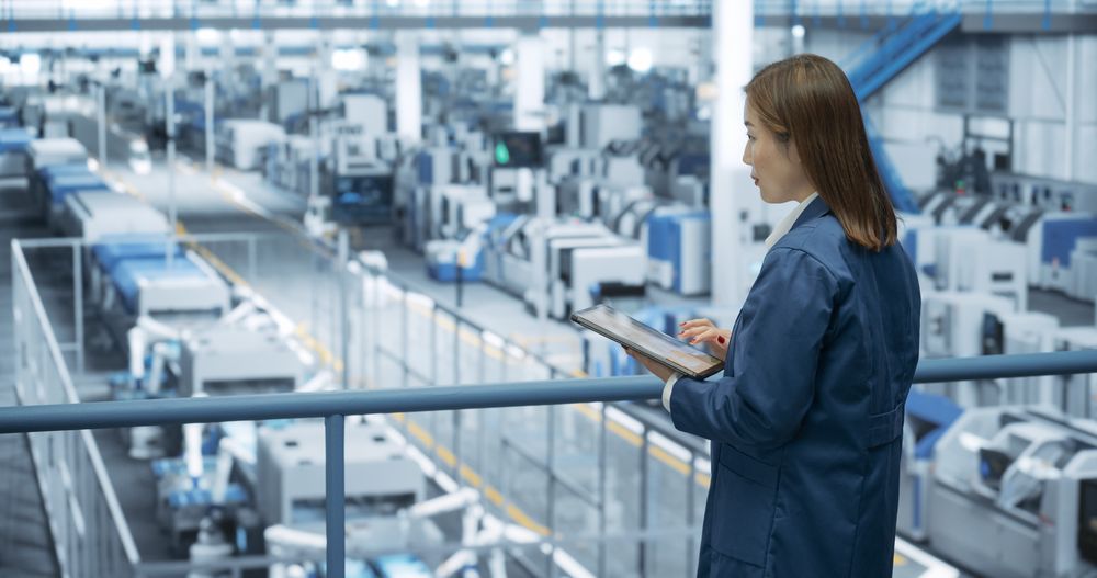Young Asian Female Engineer Standing on a Platform, Using Tablet Computer at an Electronics Factory. Machines are Undergoing Maintenance, Specialist Monitoring the Progress Through Online Software