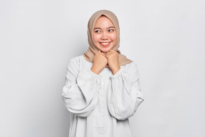 Happy Muslim woman in headscarf in light blouse with hands under her chin looking shy