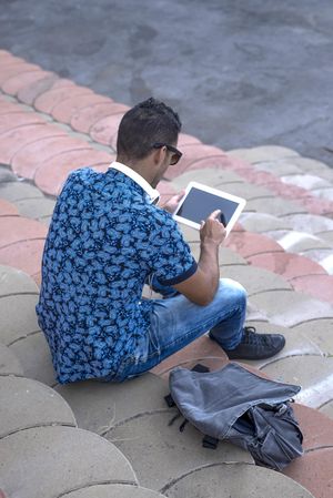Rear of male sitting on stairs on sunny day and using tablet, vertical