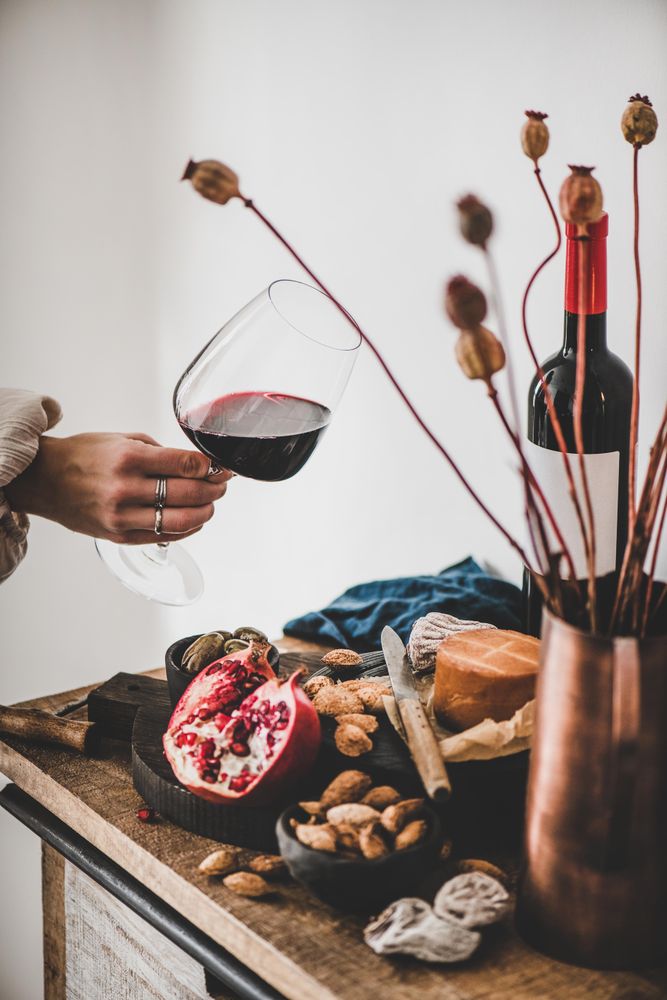 Rustic setting of women holding wine, with cheese, long candles, pomegranate, and dried poppies