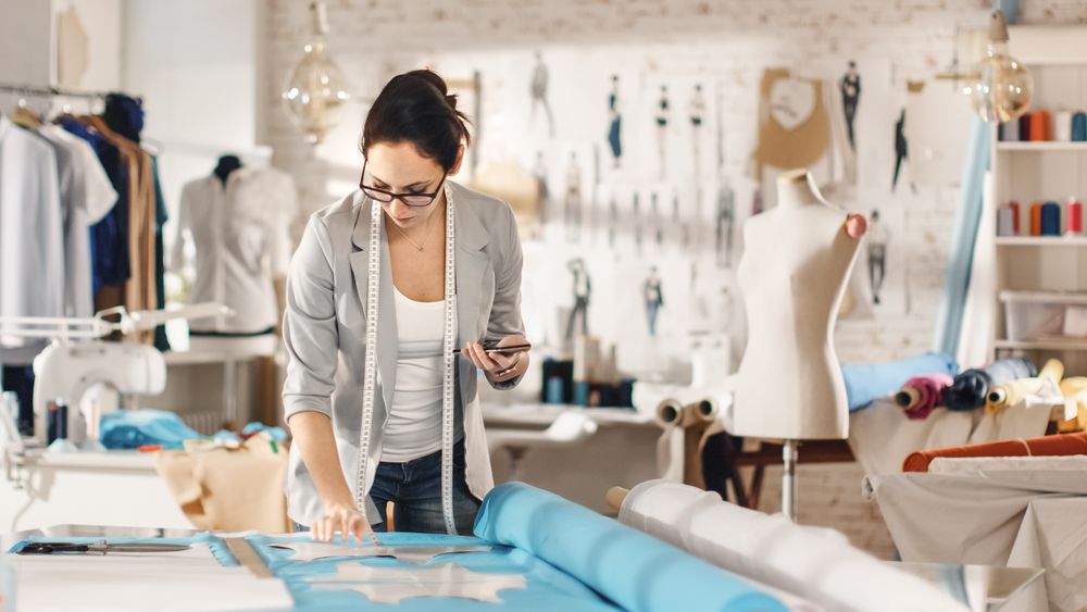 Brunette fashion, designer,  Lays Out Templates on the fabrics and Consults Her Smartphone.