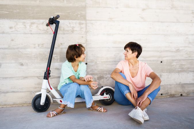 Woman and preteen girl sitting on the ground outside with a scooter