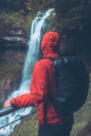 Back view of a man in red jacket with backpack standing beside fence looking at waterfall