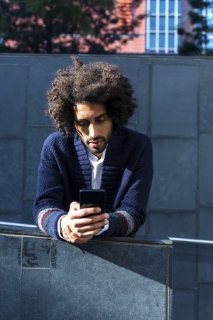Black man in cardigan leaning forward outdoors on sunny day looking at phone