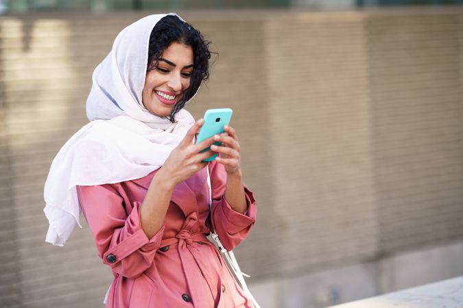 Smiling female in headscarf and pink trench coat sitting outside with her phone