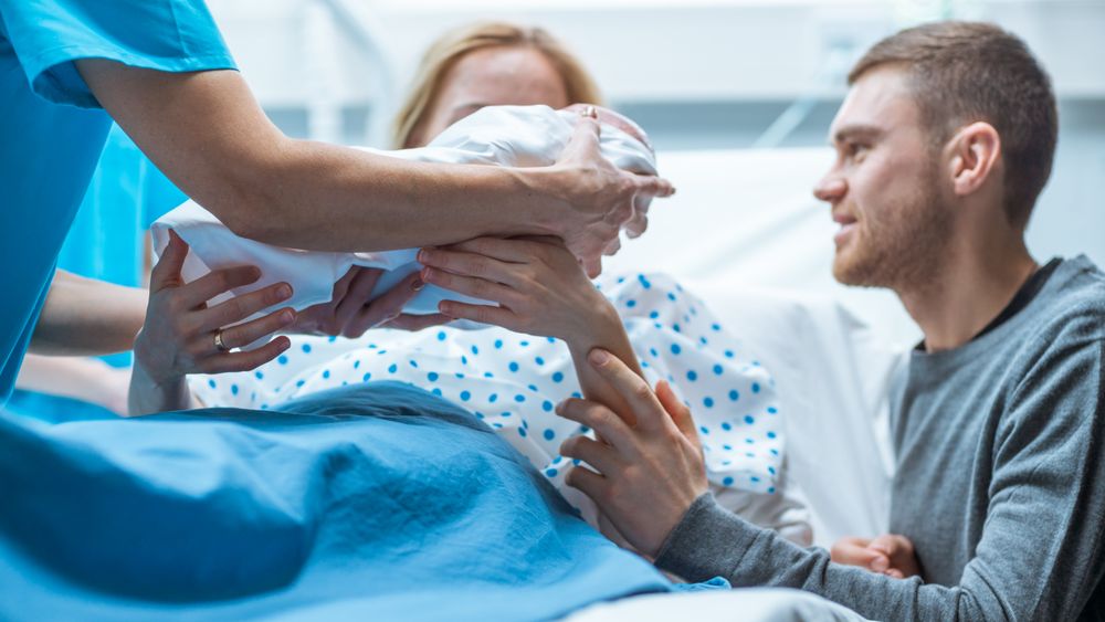 In the Hospital Midwife Gives Newborn Baby to a Mother to Hold, Supportive Father Lovingly Hugging Baby and Wife