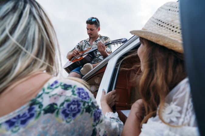 Man playing on guitar on car roof and women listening with pleasure