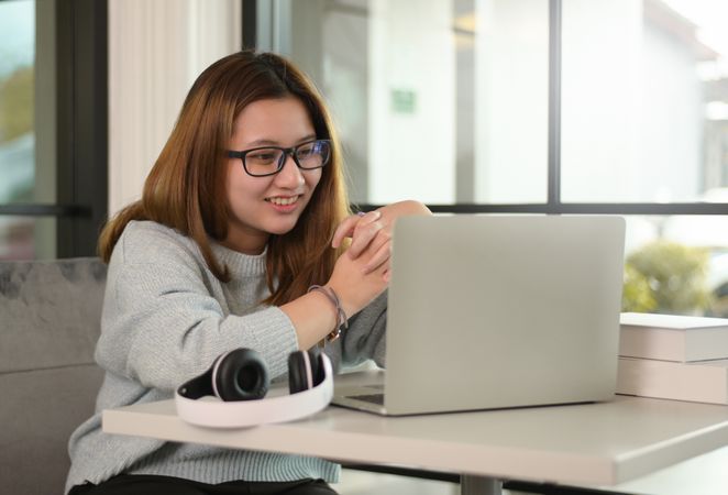 A woman wearing glasses with a laptop and headphones on the table is chatting with a smiling friend.