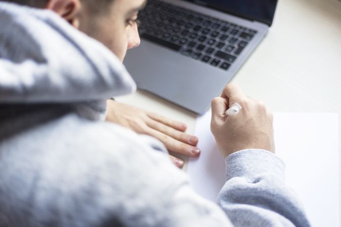 Rear view of a young man writing on paper next to laptop at home