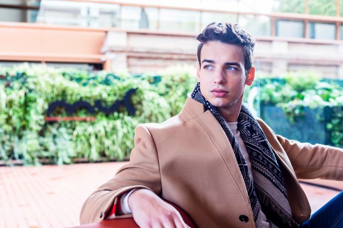 Young man wearing winter clothes looking around sitting on a leather bench outside