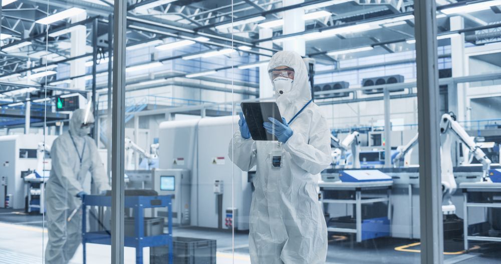 Portrait of a Female Worker Wearing Full-Body Protective Coveralls, Mask and Goggles at a Biotechnology Manufacturing Plant. Young Specialist Using a Tablet Computer, Overlooking Nanotech Production