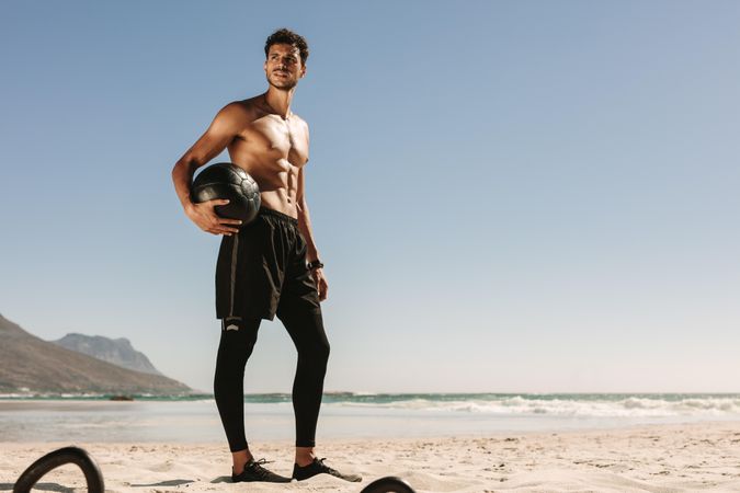 Physically fit male posing with weighted ball at a beach