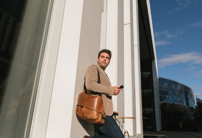 Businessman with smartphone leaning on building on city street