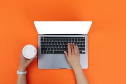 Person using laptop on orange desk with take away cup of coffee or tea with copy space